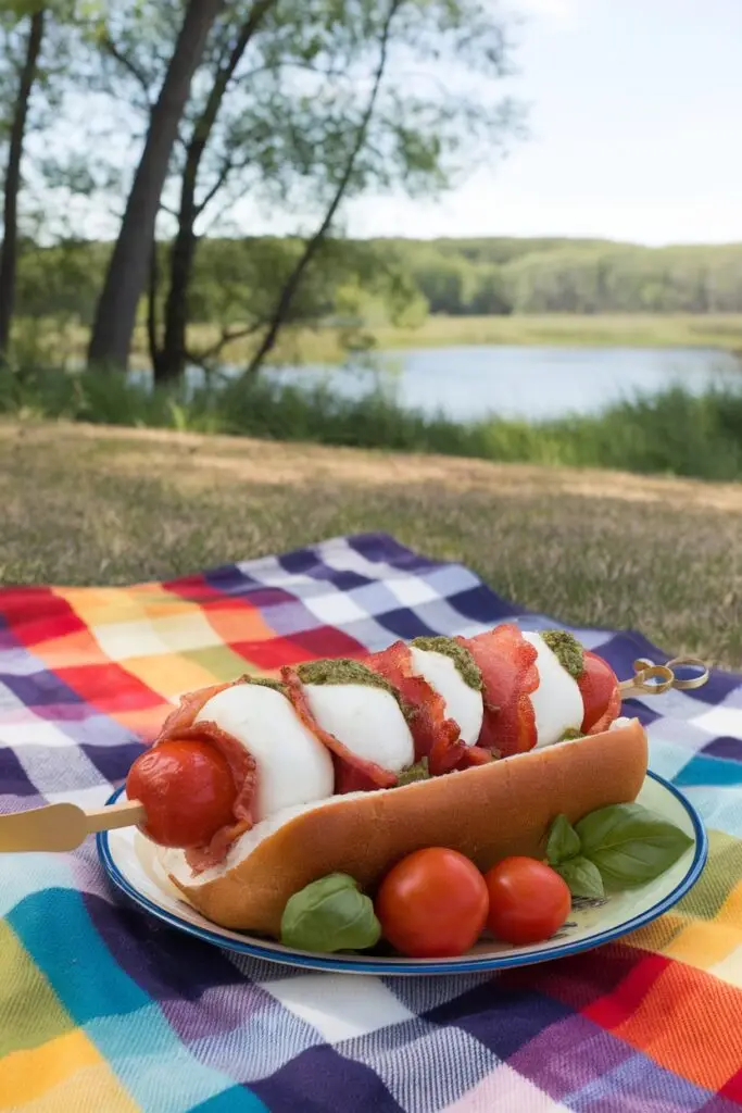 A caprese-style hot dog with mozzarella, tomatoes, and basil pesto, set on a picnic blanket by a lake.