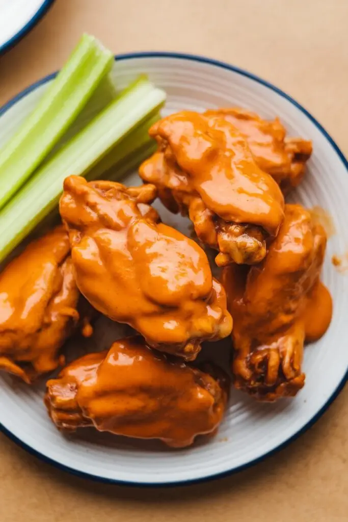 A plate of golden, crispy buffalo wings coated in a rich, spicy sauce with a side of celery.