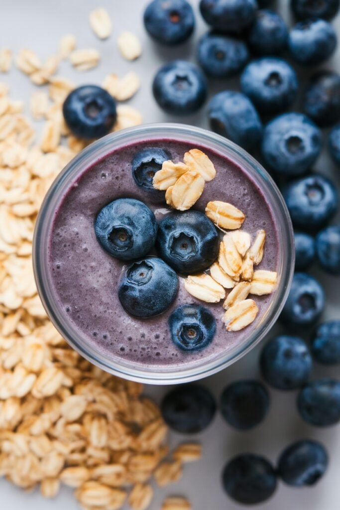 A glass of blueberry oat smoothie topped with fresh blueberries and rolled oats, surrounded by more blueberries and oats.