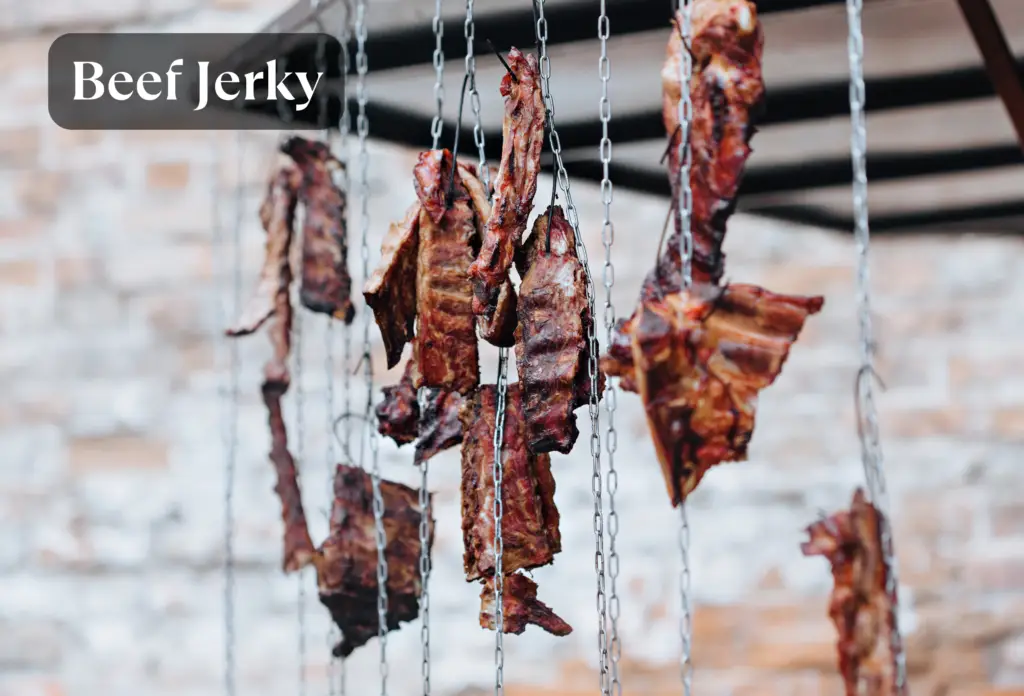 Strips of dark red beef jerky hanging from metal chains against a blurred brick wall background, with a "Beef Jerky" text overlay in the upper left corner.