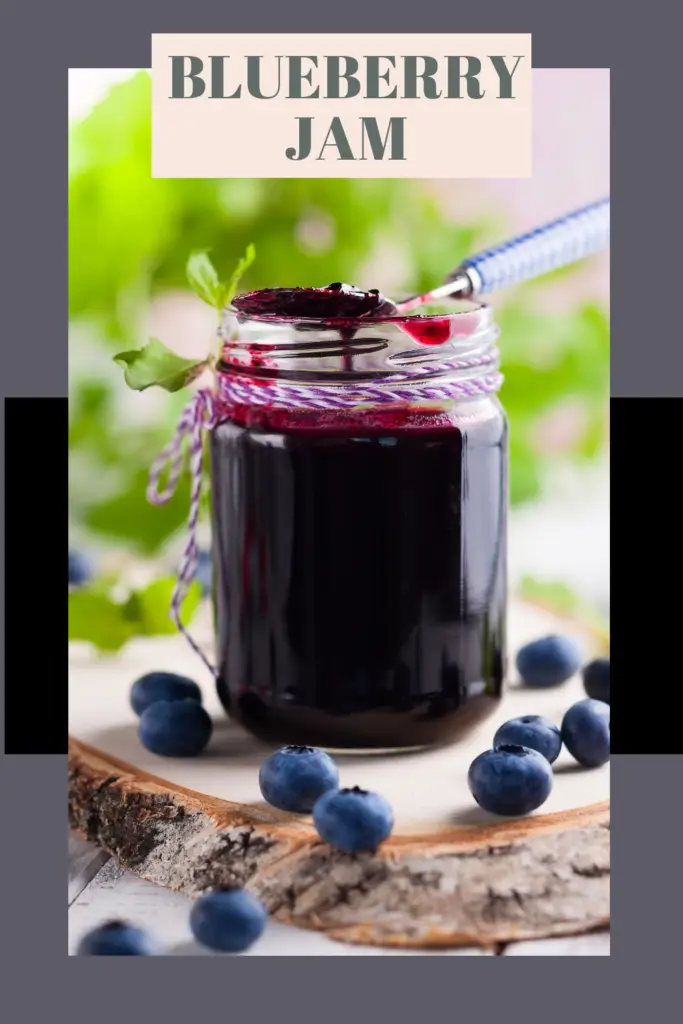 A jar of blueberry jam with a spoon, surrounded by fresh blueberries and greenery.