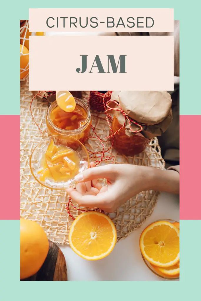 Hand holding a glass of orange jam with jars of citrus preserves, fresh orange slices, and decorative packaging on a woven mat against mint green and pink background.