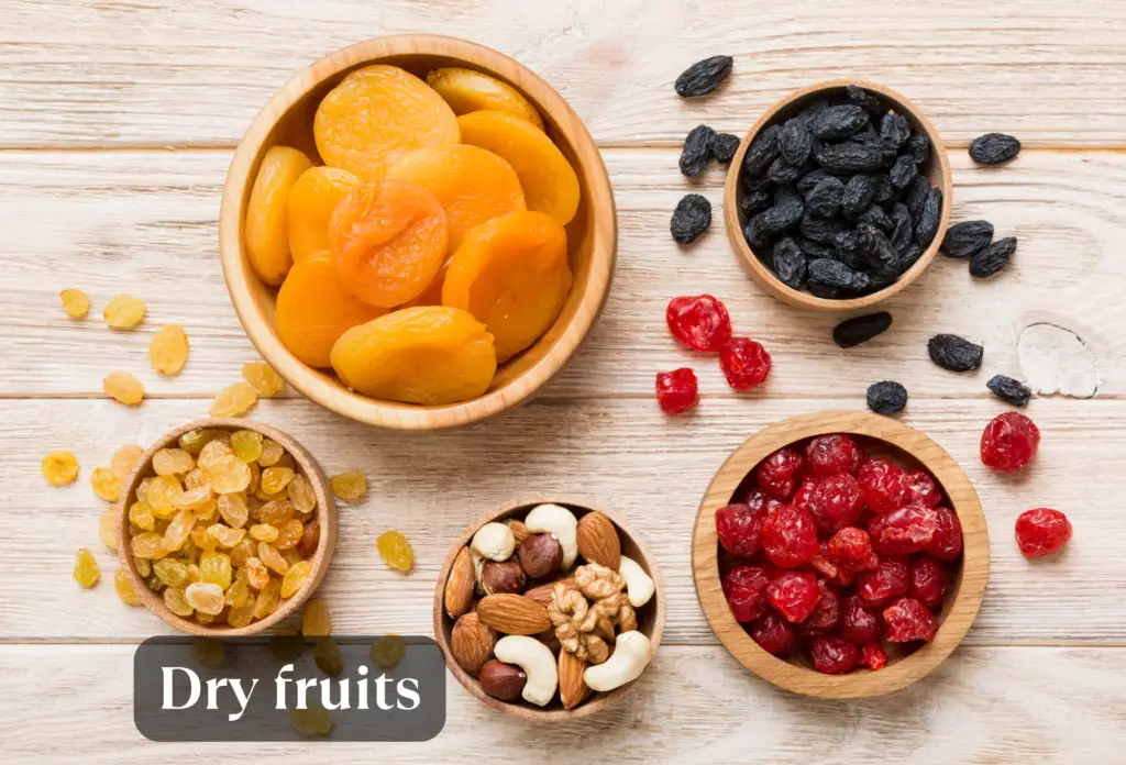 Top-down view of five wooden bowls containing various dried fruits and nuts arranged on a light wooden surface, featuring dried apricots, golden raisins, black raisins, dried cranberries, and mixed nuts, with a "Dry fruits" text overlay at the bottom.