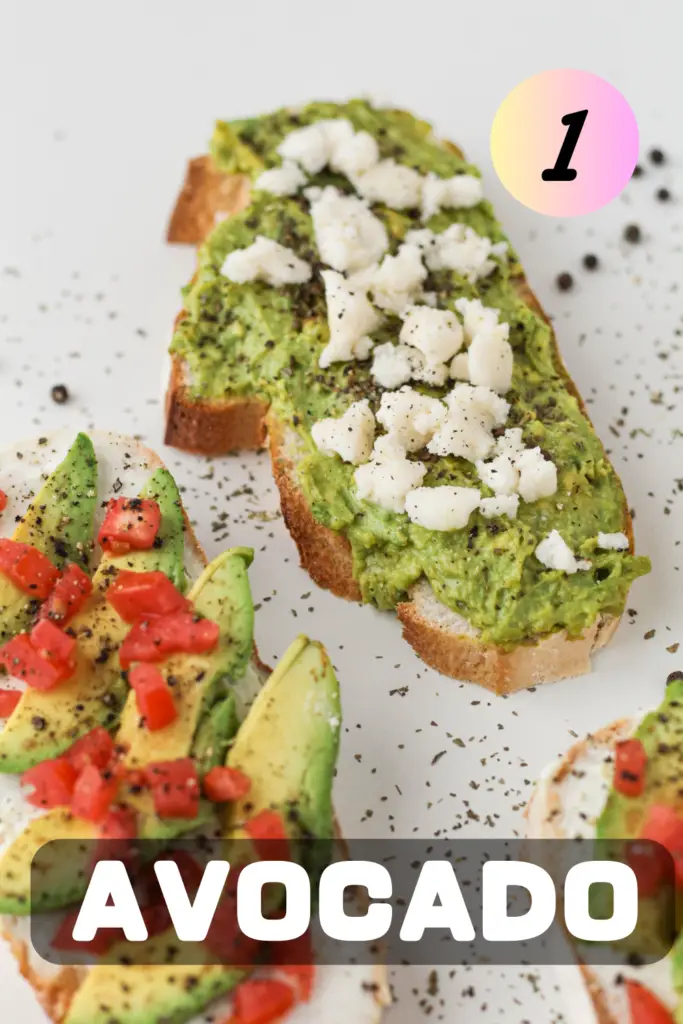 Gourmet avocado toast topped with crumbled feta cheese and black pepper on toasted bread, with additional avocado slices and diced tomatoes visible in the background on a white surface.