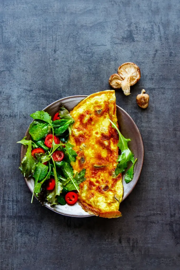 Overhead shot of a golden folded omelette on a matte brownish-gray ceramic plate with a fresh side salad of mixed greens and halved cherry tomatoes, garnished with dried mushrooms against a textured dark blue-gray background.