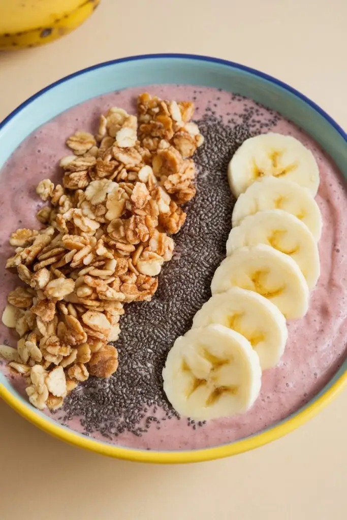 A pink smoothie bowl topped with granola, chia seeds, and sliced banana in a blue-rimmed ceramic bowl on a wooden surface.