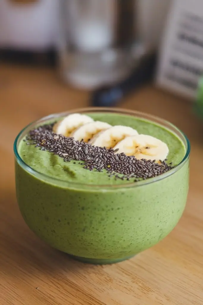 Bright green banana spinach smoothie bowl in a glass bowl, topped with sliced bananas and a stripe of black chia seeds, served on a wooden table surface