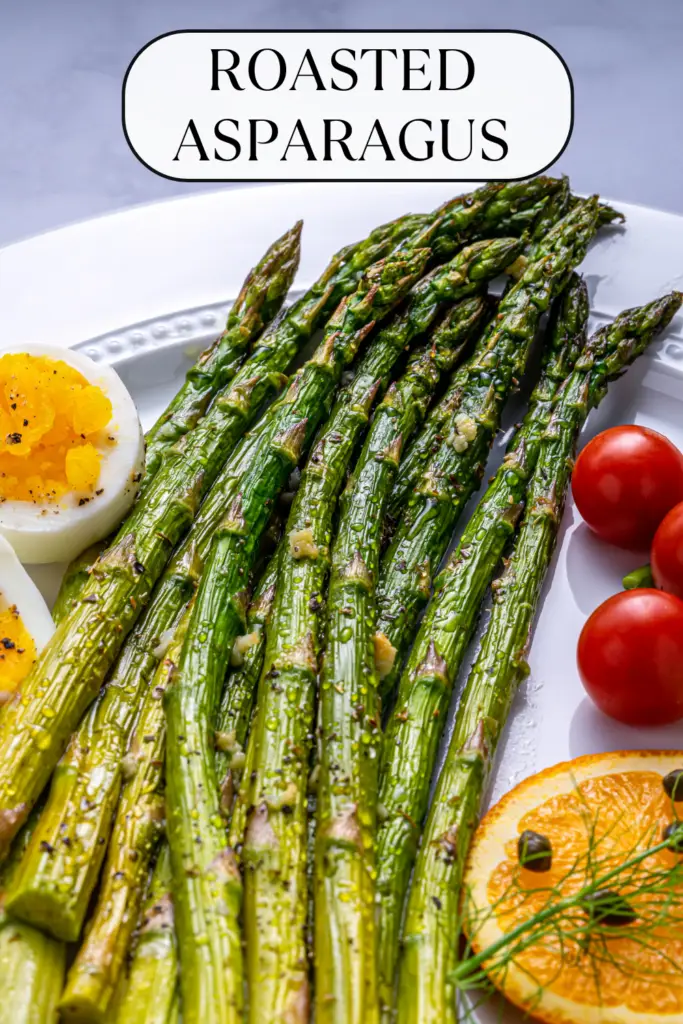 Vibrant roasted asparagus spears served with sliced boiled egg, cherry tomatoes, and orange slice on a white plate, showcasing the perfect tenderness of oven-roasted asparagus.
